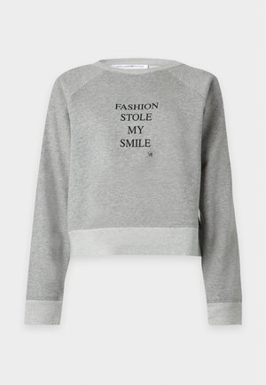 Γυναίκα που φοράει γκρι φούτερ με κείμενο "FASHION STOLE MY SMILE", μαύρη φούστα με μεγάλο σκίσιμο και μαύρα σανδάλια με λουράκια και τακούνι, στέκεται σε λευκό φόντο.