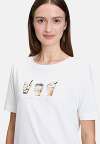 T-shirt en coton blanc avec un motif imprimé de trois boissons au café avec des pailles, accompagné du texte "coffee first" dans une police douce.