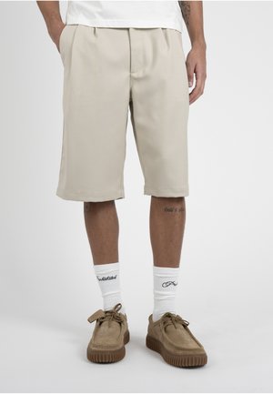 Uomo che indossa pantaloncini beige fino al ginocchio, calzini bianchi con scritte nere e scarpe marroni in camoscio con lacci su sfondo uniforme.