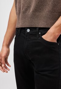 Jeans de mezclilla negra con un corte recto, diseño de cinco bolsillos y un botón metálico plateado con el logo de "Lee". Se llevan con un suéter de punto marrón.