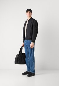 Chaqueta bomber negra con puños y cuello acanalados, camiseta beige, jeans de mezclilla azules, zapatillas negras y un bolso de duffel negro.