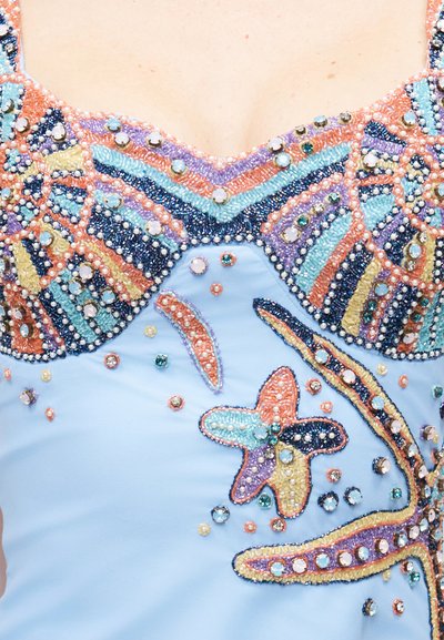 Κοντινό πλάνο ανοιχτό μπλε υφάσματος με χρωματιστά διακοσμητικά από χάντρες και πολύτιμους λίθους που σχηματίζουν γεωμετρικά σχήματα και αστερίες κατά μήκος μιας λαιμόκοψης.