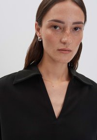 Donna con lunghi capelli castani che indossa un top nero a V con colletto e orecchini a cerchio in argento, su uno sfondo semplice e chiaro.