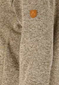 Ljusbrun jacka med strukturerad tyg och subtilt mönster, slim fit, och en liten läderlogotyp på axeln.