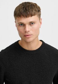 Black knit sweater with a ribbed texture and a crew neck design. Model has short hair and is wearing a subtle hoop earring.