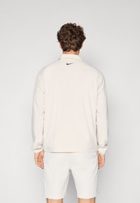 Light beige Nike jacket with long sleeves, a collar, and a small black logo at the back. Smooth texture and slightly fitted design.