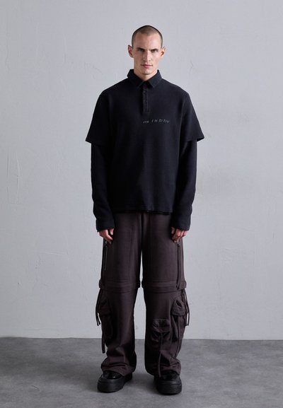 Mann mit rasiertem Kopf trägt ein geschichtetes schwarzes Langarm-Polo und lockere dunkle Cargo-Hosen mit mehreren Taschen, steht auf grauem Boden.