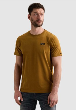 Homme aux cheveux courts et foncés avec une barbe, portant un t-shirt jaune moutarde à manches courtes et un jean foncé, regardant sur le côté sur un fond uni.