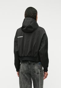 Juodas sutrumpintas bomber tipo švarkas su gaubtu, elastinėmis rankogaliais ir juosmeniu. Ant nugaros yra baltos spalvos teksto logotipas, pagamintas iš glotnaus medžiagos.