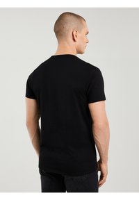 Homme aux cheveux courts portant un t-shirt noir ajusté et un jean foncé, debout de dos sur un fond gris clair.
