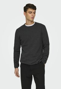 Donkergrijze langemouwen shirt met een ronde hals en textuurstof. Gecombineerd met zwarte broek, handen in de zakken, een plain witte ondershirt zichtbaar.