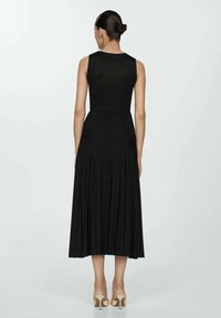 Schwarzes ärmelloses Kleid mit einer taillierten Oberteil und plissiertem Rock, aus einer strukturierten Stoffqualität, der bis zur Wade fällt, kombiniert mit nude hohen Schuhen.