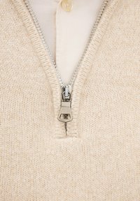 Beige gebreide ziptrui met een zilveren rits, met een hoge kraag en een crèmekleurig onderhemd met een knop.