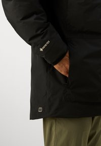 Close-up af en person iført en sort GORE-TEX jakke med hånden i lommen og olivengrønne bukser, synlige under jakken.