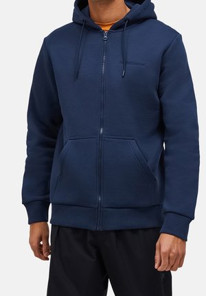 Mężczyzna ubrany w granatową bluzę z kapturem na zamek błyskawiczny, z przednimi kieszeniami i sznurkiem przy kapturze, w połączeniu z czarnymi spodniami i pomarańczową koszulką pod spodem.