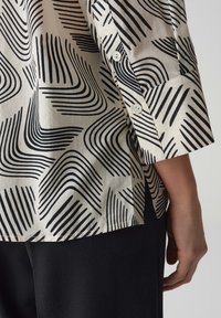 Chemise à boutons en tissu léger avec un motif abstrait de vagues noires sur fond crème. Détails visibles de boutons et de poignets.