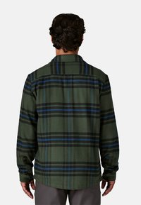 Camicetta in flanella verde a quadri con strisce nere e blu, maniche lunghe e polsini con bottoni. La vista posteriore mostra un colletto e una texture liscia.