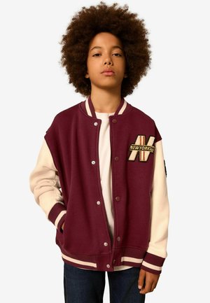 Bordeaux-rot und cremefarbene Varsity-Jacke mit Druckknöpfen, gerippten Bündchen und gesticktem "NEW YORK"-Patch auf der Vorderseite. Glatte Stoffoberfläche.