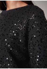 Tela negra con lentejuelas y superficie texturizada. El diseño presenta un ajuste holgado, caracterizado por un patrón intrincado de lentejuelas de varios tamaños.