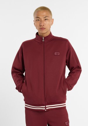 Veste zippée bordeaux en tissu doux, avec un col montant, des accents rayés blancs au niveau de l'ourlet et un petit patch logo sur la poitrine.
