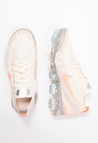 Sneakers Nike con tomaia beige in maglia, dettagli arancioni e suola traslucida con cuscinetto ad aria. Il design presenta una finitura traspirante e testurizzata.