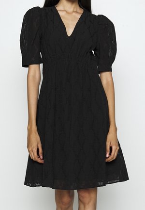 Nainen, joka on pukeutunut polvipituiseen mustaan mekkoon, jossa on puhvihihat puoli-hihoilla, kohokuvioitu timanttikuvio ja V-pääntie, seisoo yksinkertaisen taustan edessä.