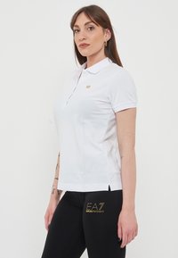 Maglietta polo bianca con colletto classico, maniche corte e un piccolo logo dorato sul lato sinistro del petto; abbinata a leggings neri con una grafica dorata lucida.
