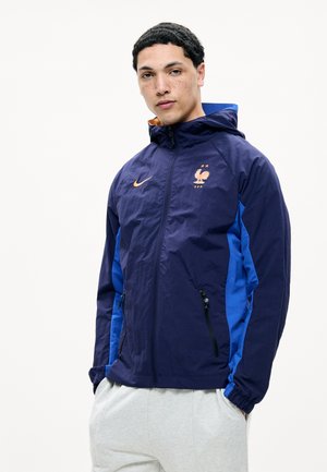 Jeune homme portant une veste à capuche bleu marine et bleu avec le logo de l'équipe de football de France et un pantalon gris clair, debout devant un fond blanc.