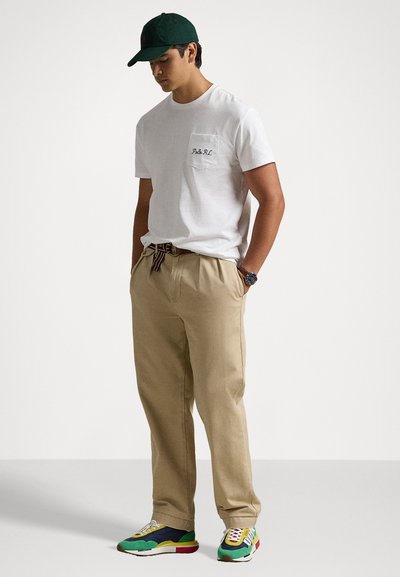 White short-sleeve t-shirt with chest pocket, beige pants, and multicolored sneakers. Accessories include a green cap and striped belt.