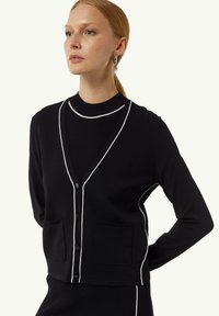 Schwarze Strickjacke mit weißen Abschlüssen, V-Ausschnitt und zwei Fronttaschen. Aus einem weichen Material gefertigt, mit tailliertem Schnitt und gerippter Textur.