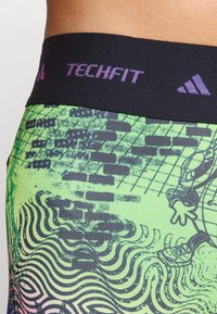 Svart midjeband med "TECHFIT" i lila text; grön tyg med abstrakta svarta mönster och vågiga linjer; slät textur.