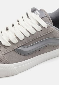 Szare zamszowe sneakersy z białymi sznurowadłami, gumową podeszwą, szarym skórzanym akcentem i perforowanymi detalami po bokach.