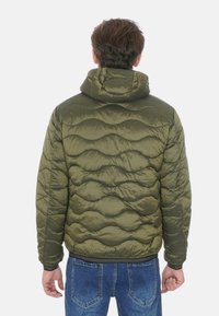 Giacca a vento imbottita con cappuccio in verde oliva, caratterizzata da un motivo trapuntato a onde, polsini elasticizzati e orlo aderente. Indossata sopra jeans blu.