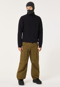 Black softshell jacket with zippered pockets, paired with loose olive cargo pants featuring multiple pockets and black boots. Complete with a knitted balaclava.