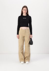 Top nero a maniche lunghe cropped con logo "HUGO", abbinato a pantaloni beige larghi con dettagli di cucitura, completato da sneakers bianche e una piccola borsa nera.