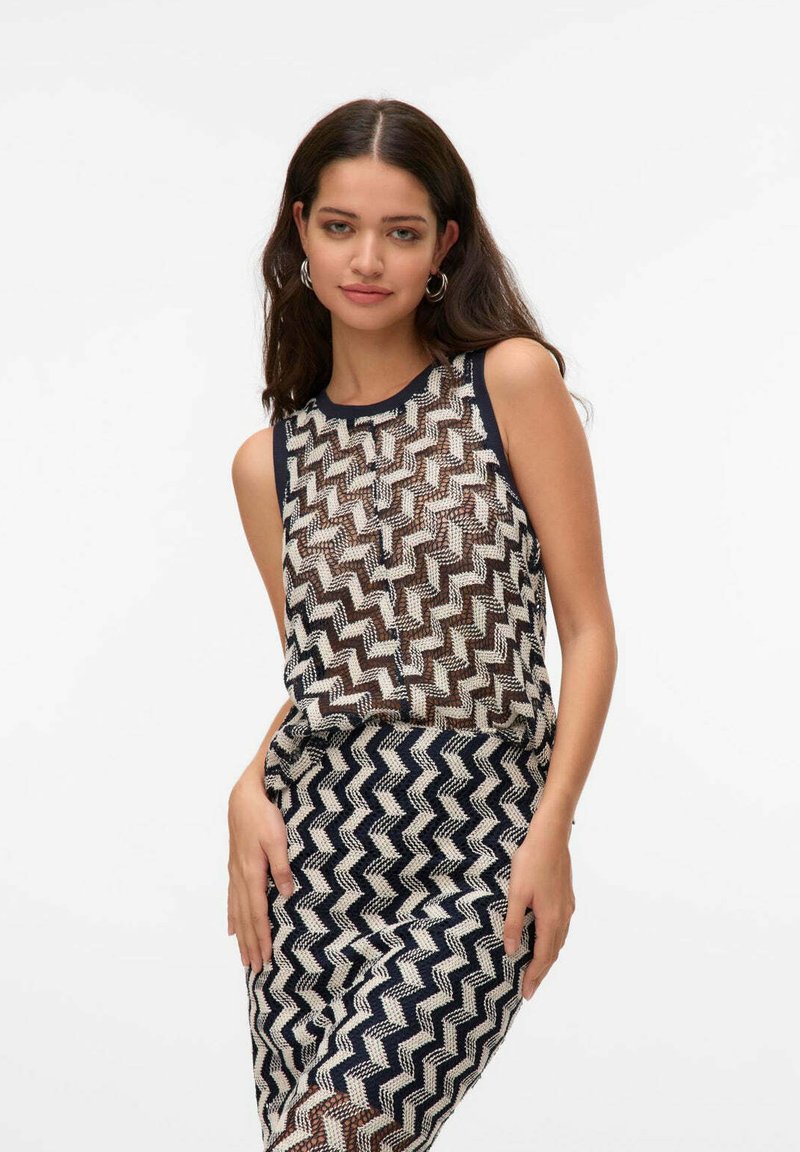 Donna con capelli lunghi e scuri indossa un vestito senza maniche a righe zigzag bianche e nere, in piedi contro uno sfondo chiaro e semplice, con le mani appoggiate sulla gonna.