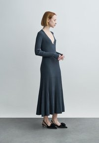 Dark gray ribbed dress with a deep V-neckline and long sleeves, flaring out at the hem. Paired with black shoes featuring floral accents.