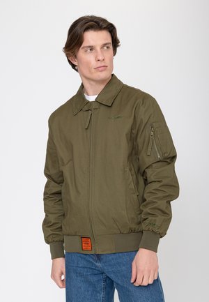 Jonge man draagt een olijfgroene bomberjas met ritszakken en blauwe spijkerbroek, kijkt zijwaarts tegen een effen achtergrond.