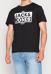 Černé bavlněné tričko s velkým bílým potiskem "JACK & JONES" na hrudi, s kulatým výstřihem a krátkými rukávy.