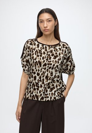 Vrouw draagt een losse korte mouwen top met luipaardprint en donkerbruine broek, staat met één hand in haar zak op een witte achtergrond.