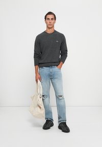 Grå stickad tröja med rund halsringning, ljusblå slitna jeans och svarta sneakers. Håller en beige tote-väska. Minimalistisk bakgrund.