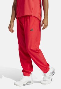 Red athletic pants with an elastic waistband, tapered legs, and a reflective gray Adidas logo on the side, paired with white sneakers.