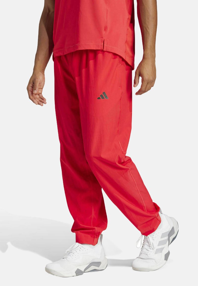 Red athletic pants with an elastic waistband, tapered legs, and a reflective gray Adidas logo on the side, paired with white sneakers.