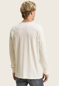 Blonder Mann mit lockigem Haar, der ein einfaches, langärmliges cremefarbenes Hemd und graue Jeans trägt und mit dem Rücken zu einem hellen Hintergrund steht.