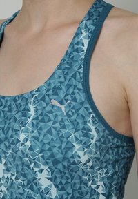Top sportivo blu decorato con motivi geometrici, spalline sottili e un grande logo bianco Puma sul petto.