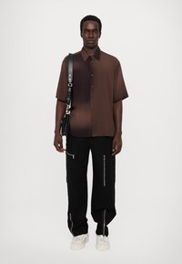 Rövidujjú barna átmeneti ing gombokkal, fekete nadrággal, amely ezüst cipzáras zsebekkel rendelkezik, és fehér sportcipővel van párosítva.