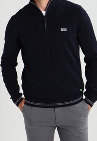 Marinblå tröja med dragkedja vid kragen, ribbade ärmslut och nederkant. Har en liten vit logotyp på bröstet. Tillverkad av ett mjukt, texturerat tyg.