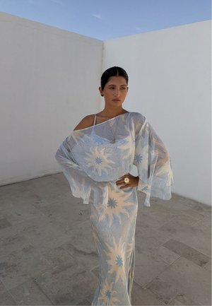 Femme aux cheveux foncés dans une robe fluide bleue et beige à motif soleil, épaules dénudées, debout avec une main sur la hanche contre des murs blancs et un sol carrelé.