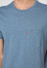 Homme portant un T-shirt bleu à col rond avec une poche poitrine et une petite étiquette rouge avec le logo Levi's sur la poche.