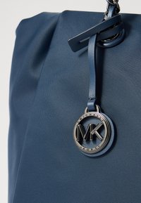 Plava najlon torba s preklopnim gornjim dijelom, s srebrnim okruglim logotipom i kožnim detaljima. Glatka tekstura, minimalistički stil.